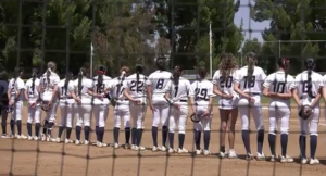 softball players in white uniforms stands shoulder to shoulder on the field during a pregame moment, seen through a protective net with trees and the outfield fence in the backgroun