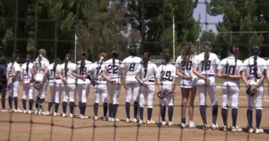 softball players in white uniforms stands shoulder to shoulder on the field during a pregame moment, seen through a protective net with trees and the outfield fence in the backgroun