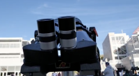 A close-up view of a telescope’s dual eyepieces points toward the sky, set up outdoors on a college campus with buildings and people gathered in the background for an astronomy event.