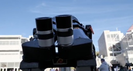 A close-up view of a telescope’s dual eyepieces points toward the sky, set up outdoors on a college campus with buildings and people gathered in the background for an astronomy event.