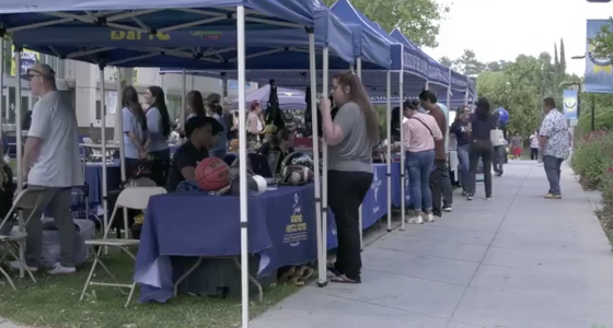Students and staff gather under a row of blue canopy tents on campus, browsing booths and talking with program representatives during an outdoor college event.