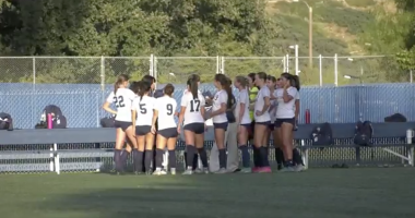 COC Wonen's soccer team wearing white jerseys and dark shorts huddles together on a green turf field during a sunny day.