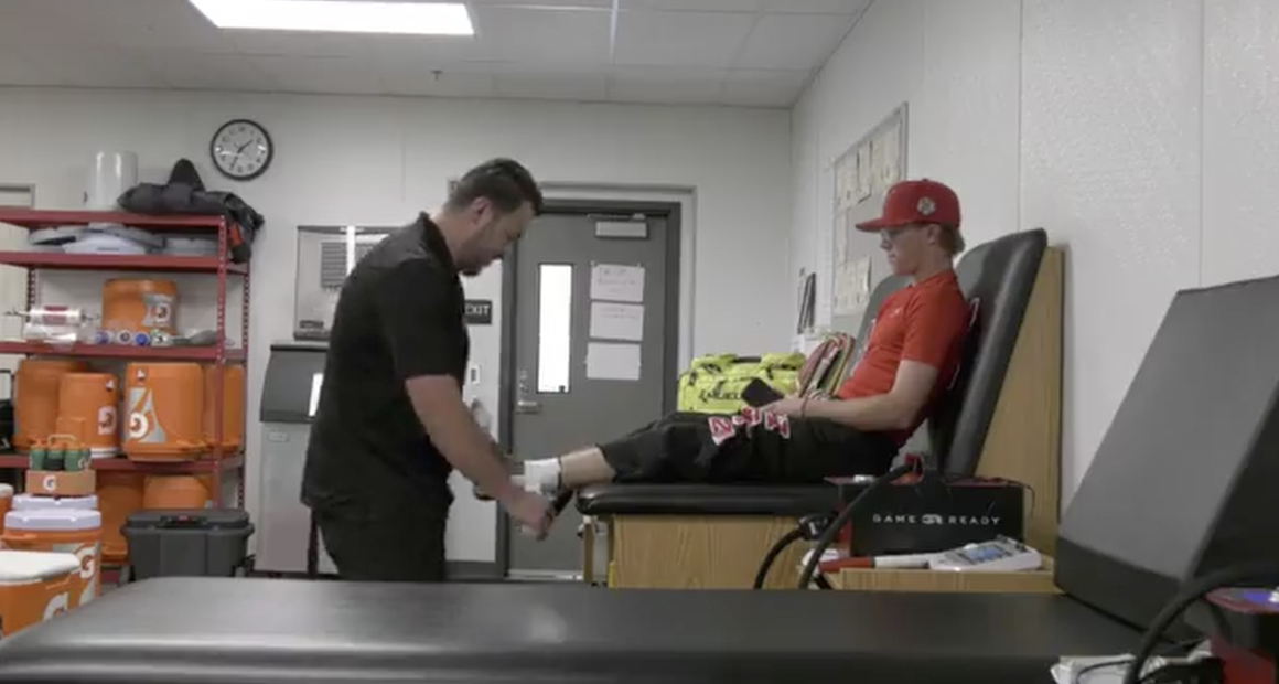 A sports trainer evaluates a young athlete’s leg on a treatment table in an athletic training room, surrounded by medical equipment and supplies.