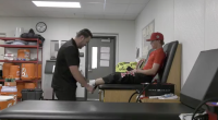 A sports trainer evaluates a young athlete’s leg on a treatment table in an athletic training room, surrounded by medical equipment and supplies.
