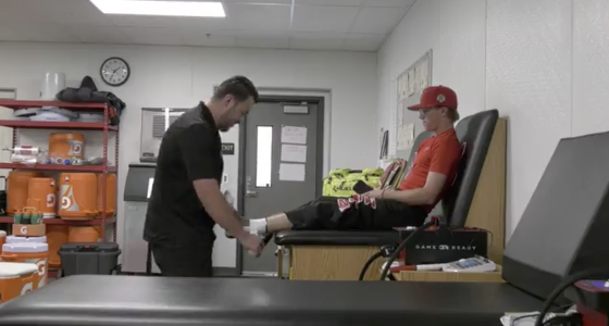 A sports trainer evaluates a young athlete’s leg on a treatment table in an athletic training room, surrounded by medical equipment and supplies.